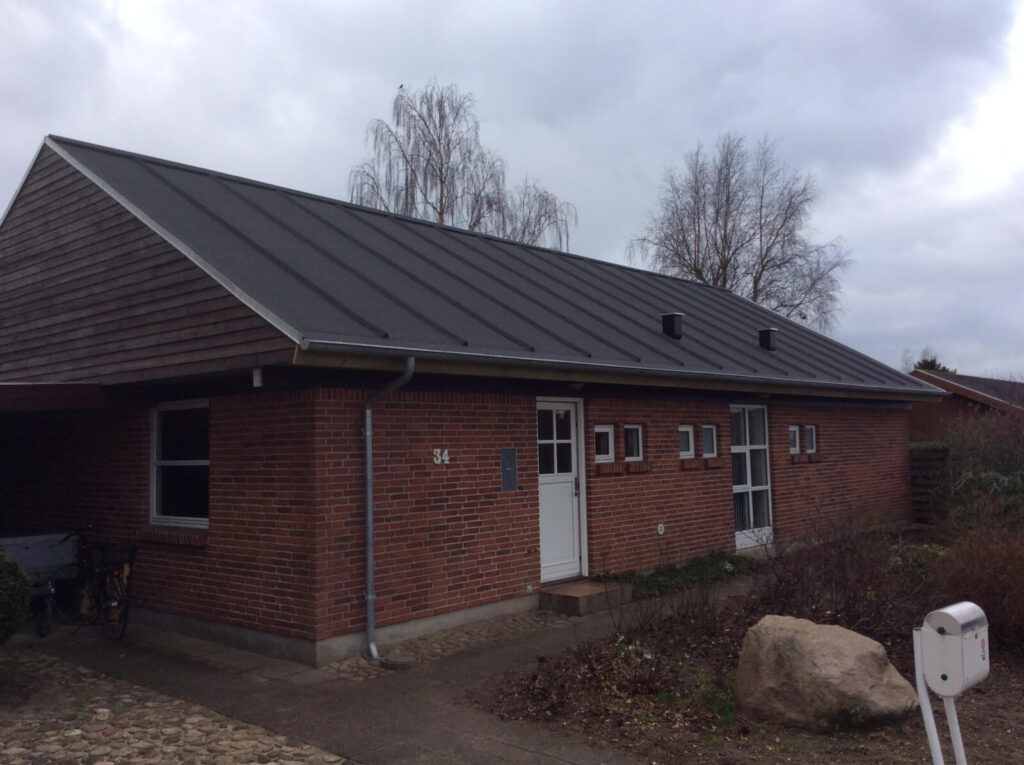En muret bungalow med et sort tag og hvide tagrender under en grå himmel med træer i baggrunden.